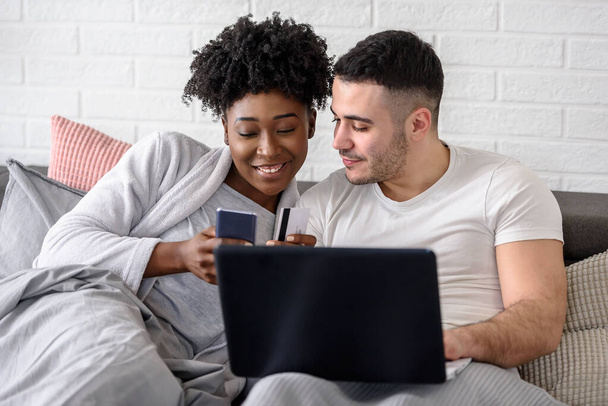 Lächelndes Mischlingspaar liegt mit Laptop im heimischen Schlafzimmer auf dem Bett  - Foto, Bild