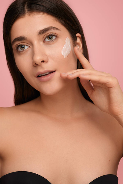 Young female model with bare shoulders applying moisturizing cream on face standing against pink background - Фото, зображення