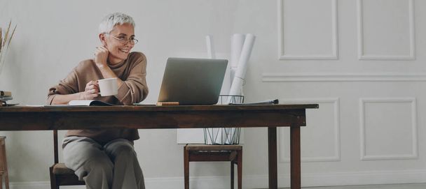 Happy senior businesswoman enjoying hot drink while using laptop at her working place - Foto, Bild