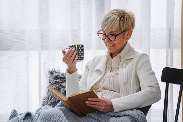 mulher sênior ler livro e beber chá em casa
 - Foto, Imagem