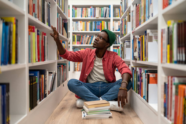 Zelfstudie. Jonge Afro-Amerikaanse man zit op de vloer in de bibliotheek tussen de boekenplanken, tijd doorbrengen in de boekhandel, op zoek naar boek, zwarte mannelijke student die zich voorbereidt op college test. Wereldboekendag - Foto, afbeelding