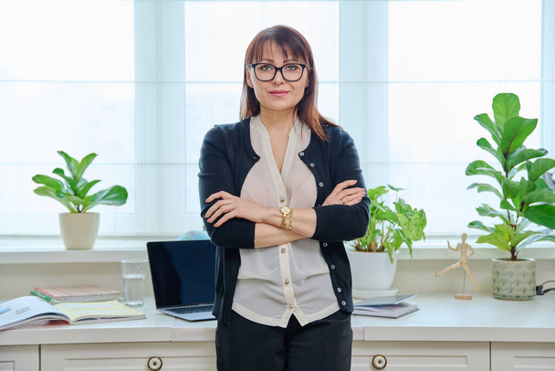 Portrait of confident mature woman in home office. Middle-aged female looking at camera with crossed arms. Freelance, remote business, online teacher psychologist accountant legal, internet service - Valokuva, kuva