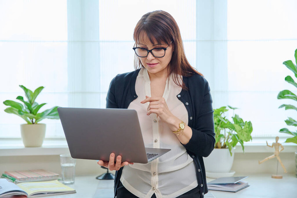 Portrait of middle aged woman in glasses looking at screen, female using laptop in front of window in office. Freelance, remote business, online teacher psychologist accountant legal, internet service - 写真・画像