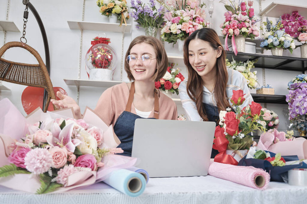 Two young beautiful female florist partners discuss flora bunch decoration, online purchase orders and website arrangement for business, happy work in colorful flower shop store, and e-commerce SME. - Fotoğraf, Görsel