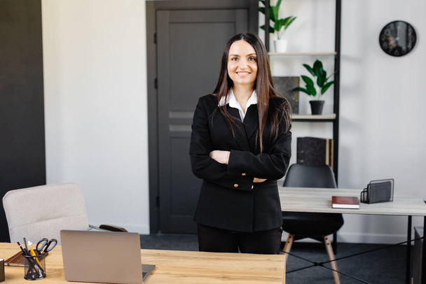 Πορτραίτο μιας επιχειρηματία σε ένα γραφείο. Μια όμορφη νεαρή γυναίκα στέκεται στο γραφείο κοντά στο χώρο εργασίας της με τα χέρια σταυρωμένα μπροστά της.. - Φωτογραφία, εικόνα