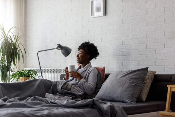 afro-americana tendo descanso em casa, deitada na cama, bebendo chá - Foto, Imagem