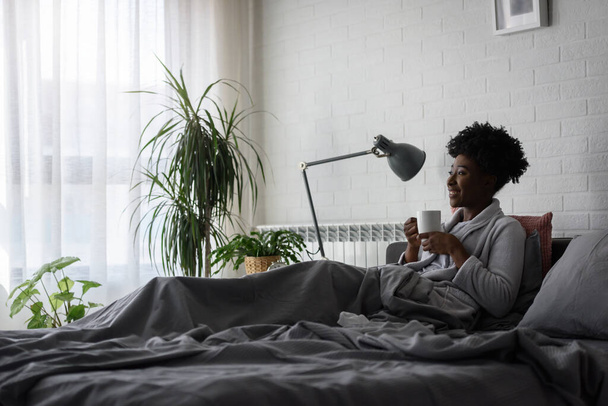 african american woman having rest at home, lying on bed, drinking tea - 写真・画像