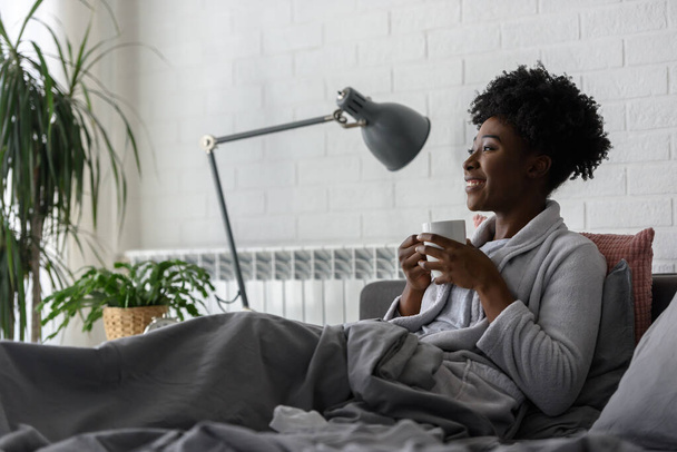 Afrikaans amerikaanse vrouw die rust thuis, liggend op bed, thee drinken - Foto, afbeelding