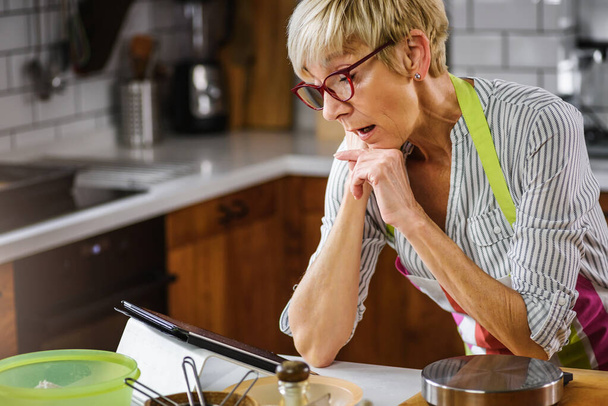 Femme âgée qui cuisine dans la cuisine de la maison. Utilisation d'un ordinateur tablette pour trouver la recette. Femme mûre cuisine avec l'aide d'Internet. Technologie moderne pour les personnes âgées. - Photo, image