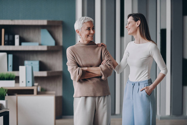 Two businesswomen looking at each other and smiling while standing in office together - Fotoğraf, Görsel