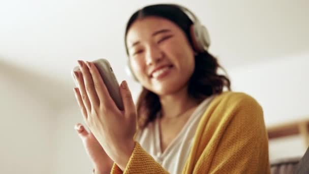 Manos de mujer, auriculares y teléfono en una sala de estar con la felicidad de la música y mensajes de texto. Inicio, sofá salón y la sonrisa de una mujer asiática y gen z sensación de estudiante relajarse de audio y podcast web. - Imágenes, Vídeo