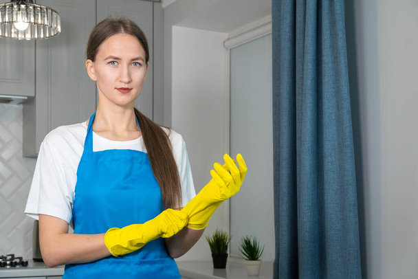Young beautiful woman in uniform puts on yellow rubber gloves for cleaning the room and looks into the camera, copy space. Cleaning concept - Foto, Imagem
