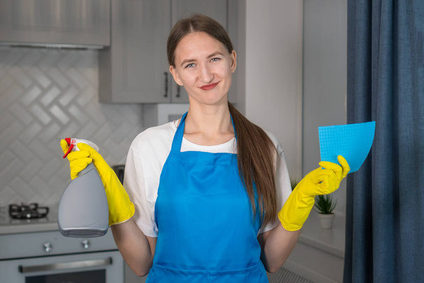 Beautiful young woman in uniform holds a cleaning agent and a microfiber cloth in her hands and looks at the camera. She is unhappy, tired, very depressed. She hates her job cleaning the apartment. - Фото, изображение