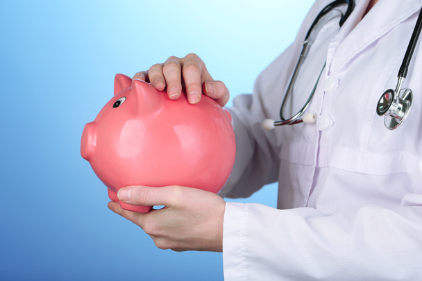 Doctor with pink piggy bank on blue background, close up - Foto, Imagen