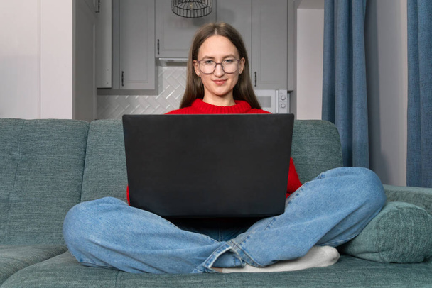 Attractive young woman with glasses is working on a laptop on the couch. Beautiful smiling girl in a red sweater makes purchases or communicates on a social network online, watches movies, freelancer. - Photo, Image