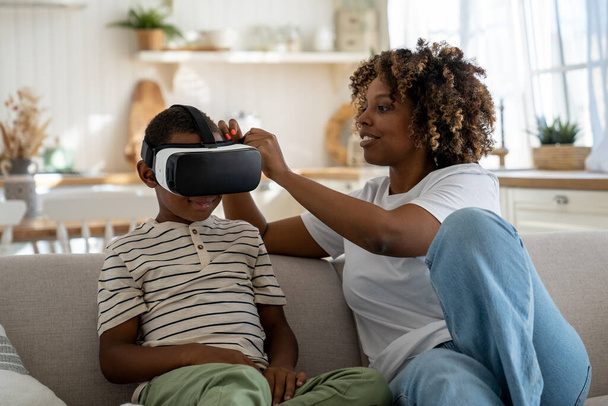 African American modern mom helping little boy son to put on virtual reality headset while enjoying leisure together on weekend, mother introducing kid with awesome technology of VR gaming - Fotó, kép