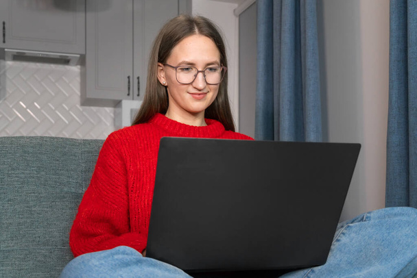 Attractive young woman with glasses is working on a laptop on the couch. Beautiful smiling girl in a red sweater makes purchases or communicates on a social network online, watches movies, freelancer. - Foto, afbeelding