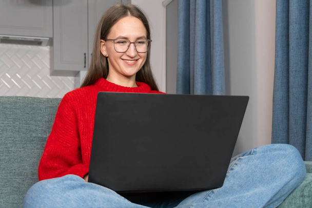Happy young woman with glasses is resting on the couch and using a laptop.An attractive girl is sitting on the sofa in the living room with her laptop while browsing the Internet.Woman in red sweater. - Foto, afbeelding
