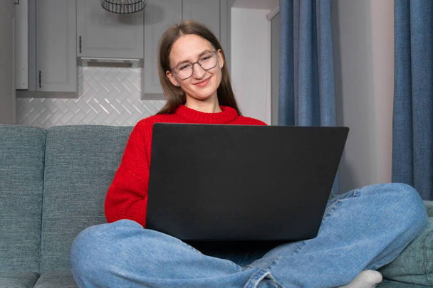 Happy smiling young woman in a red sweater,glasses,sitting on the couch,using a laptop in the living room at home,watching a funny video,learning a language,video call,mother working online,freelancer - Фото, изображение