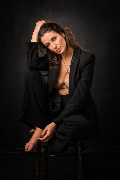 Full length of an attractive mid aged woman with toothy smile wearing black blazer and pants sitting against at isolated dark background. Copy space.  - Foto, Bild