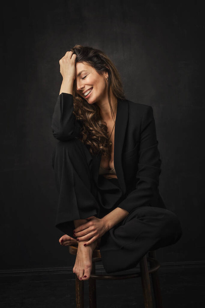 Full length of an attractive mid aged woman with toothy smile wearing black blazer and pants sitting against at isolated dark background. Copy space.  - Photo, image