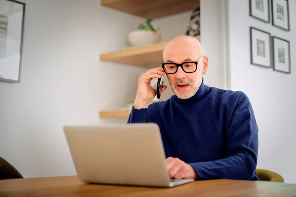 Surprised mid aged man making a call and using laptop while working from home. Confident male sitting in the living room and having a call. Home office. - 写真・画像