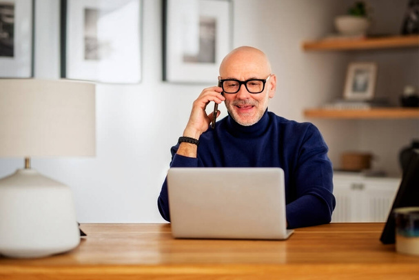 Happy middle aged man making a call and using laptop while working from home. Confident male sitting in the living room and having a call. Home office. - Foto, Imagen