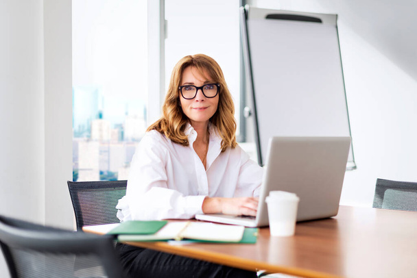 Smiling mid aged businesswoman sitting in a modern office and using laptop for work. Blond haired professional woman wearing glasses and white shirt.  - Photo, image