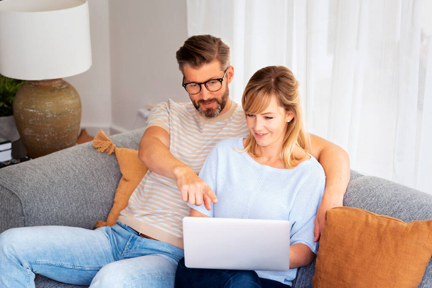 Shot of a couple using a laptop while relaxing on the couch at home. Handsome man and beautiful woman sitting together and browsing on the internet. Man showing something on computers screen.   - Photo, Image