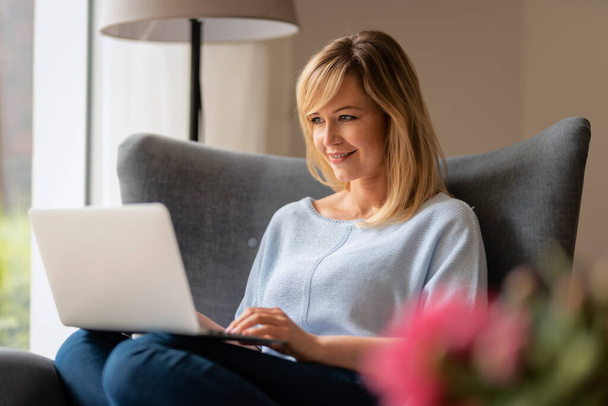 Shot of an attractive middle aged woman sitting in an armchair while using laptop. Beautiful female wearing casual clothes. Home office. - Foto, Imagen