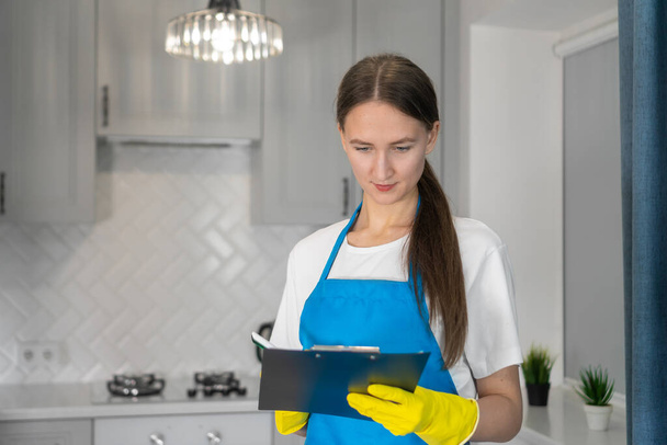Young beautiful professional cleaning woman in an apron from a cleaning company checks the work done using a checklist on a paper planter or accepts an order to perform house cleaning work - Foto, Imagem