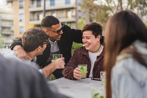 Een diverse groep vrienden, waaronder een niet-binaire plus size persoon en een blanke man, genieten van mojitos in een outdoor pub als ze gelukkig begroeten een mixed-race vriend met een knuffel. - Foto, afbeelding