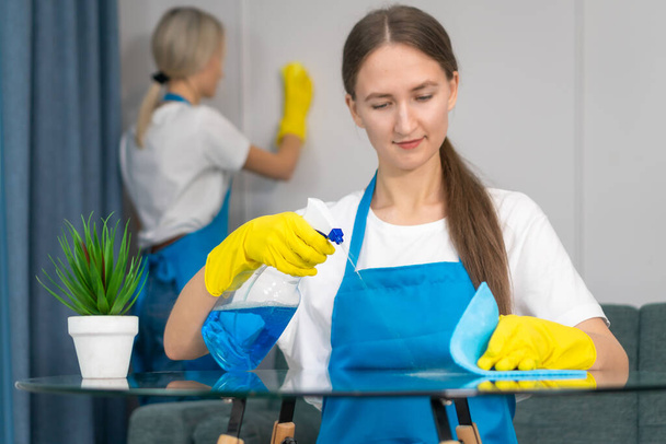 Close up staff of the cleaning service with professional equipment is engaged in cleaning the house, hygiene. Young woman in uniform wipes the table with a cleaning agent, microfiber cloths. - Фото, изображение