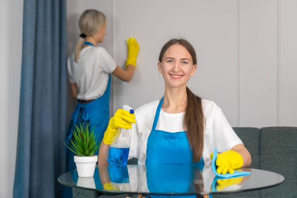 専門的な設備を備えた清掃サービスのスタッフは、家、衛生の清掃に従事しています。制服を着た若い女性は、洗浄剤、マイクロファイバークロスでテーブルを拭く. - 写真・画像