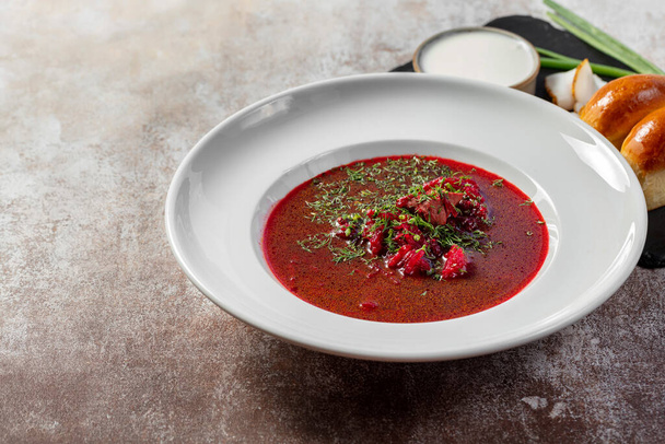 Ukrainian borscht soup with garlic donuts, green onions and bacon, next to it is a bowl of sour cream. The soup is poured into a light ceramic plate. Dishes stand on a light background. - Photo, Image