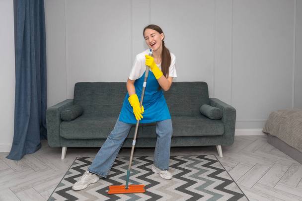 House cleaning with fun. Happy young housewife singing her favorite song while cleaning, using mop as microphone and guitar, enjoying her homework. Young woman dancing and cleaning in living room. - Fotografie, Obrázek