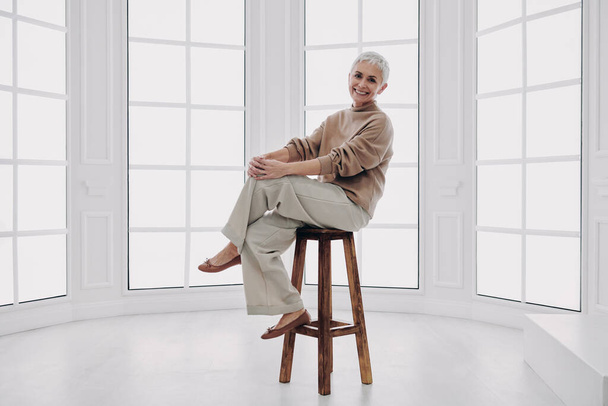 Happy senior woman looking at camera while sitting on the chair against a window - Фото, изображение