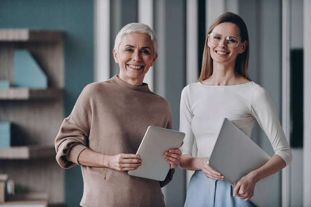 Two confident businesswomen holding gadgets and smiling while standing in the office together - Photo, Image