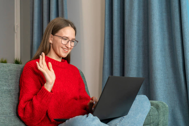 Young smiling woman in glasses, a red sweater waves her hand while sitting at a laptop on the couch. Attractive girl greets in video chat, with colleagues, friends, online communication - Photo, image