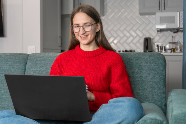 Happy smiling young woman in a red sweater,glasses,sitting on the couch,using a laptop in the living room at home,watching a funny video,learning a language,video call,mother working online,freelancer - Foto, afbeelding