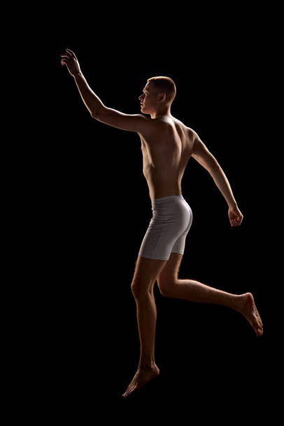 Male body in motion. Portrait of young attractive redhead man wearing underwear and jumping in unusual posture over dark background. Concept of futurism, surrealism, fashion, cyber, beauty, art, ad - Фото, изображение