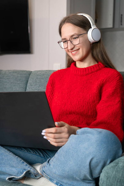 Young woman in a red sweater, jeans, white headphones is surfing the net on the couch at home. Girl working in a home office. Freelancer using a laptop and the Internet. Workplace in the living room - 写真・画像