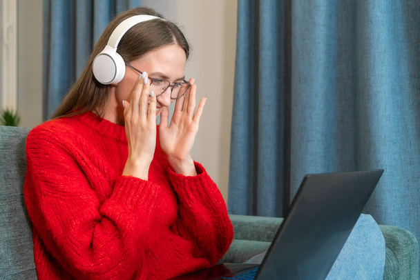 Χαριτωμένη νεαρή χαμογελαστή καστανή γυναίκα σε ένα κόκκινο πουλόβερ, λευκά ακουστικά προσαρμόζει τα γυαλιά της, ενώ κοιτάζοντας ένα φορητό υπολογιστή, ενώ κάθεται στον καναπέ στο σαλόνι. Απομακρυσμένη εργασία, ελεύθερος επαγγελματίας - Φωτογραφία, εικόνα