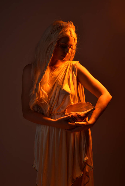portret van mooie blonde vrouw dragen van een fantasie godin toga kostuum met kristallen kroon.Backlit een gloeiende geïsoleerde studio achtergrond met oranje silhouet verlichting. - Foto, afbeelding