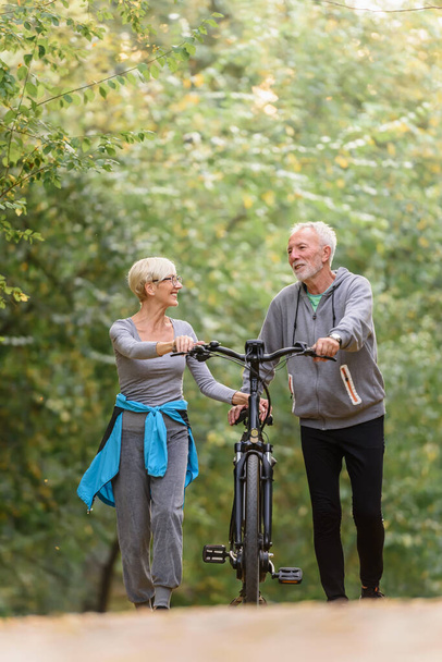 Vrolijk actief seniorenpaar met fiets wandelen door park samen. Perfecte activiteiten voor ouderen. - Foto, afbeelding