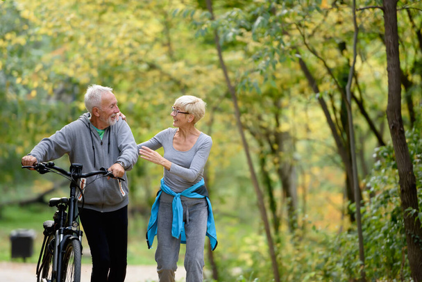 Веселая активная пожилая пара с велосипедом, прогуливающаяся по парку вместе. Идеальные занятия для пожилых людей. - Фото, изображение