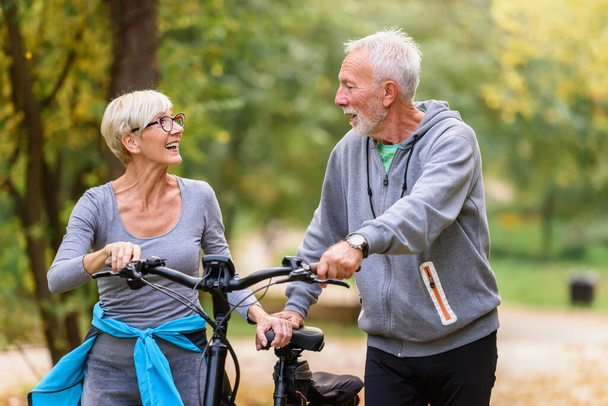 Fröhliches rühriges Seniorenpaar mit Fahrrad spaziert gemeinsam durch den Park. Perfekte Aktivitäten für ältere Menschen. - Foto, Bild