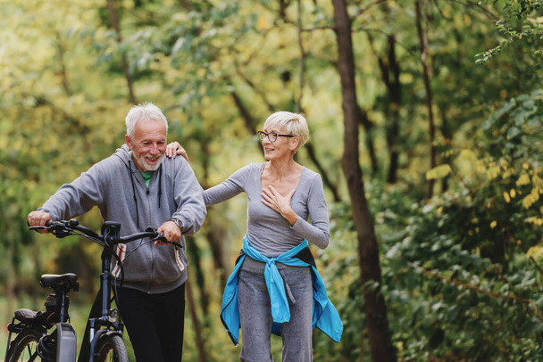 Fröhliches rühriges Seniorenpaar mit Fahrrad spaziert gemeinsam durch den Park. Perfekte Aktivitäten für ältere Menschen. - Foto, Bild