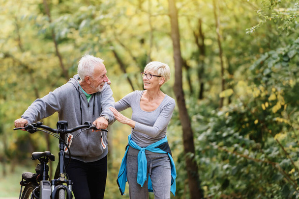 Χαρούμενο ενεργό ζευγάρι ηλικιωμένων με ποδήλατο περπατώντας μέσα από το πάρκο μαζί. Ιδανικές δραστηριότητες για ηλικιωμένους. - Φωτογραφία, εικόνα