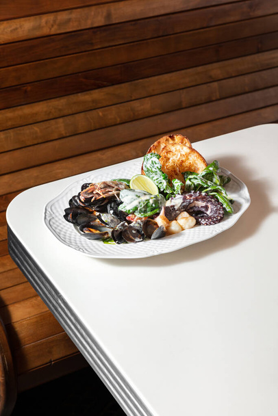 Assorted seafood from scallops, mussels, langoustines, octopus with lettuce in a creamy sauce with two toasts. Food lies in a metal plate on a light background. - Photo, Image